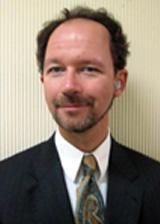 Carleton looks at the camera wearing a white collared shirt and brown coat.