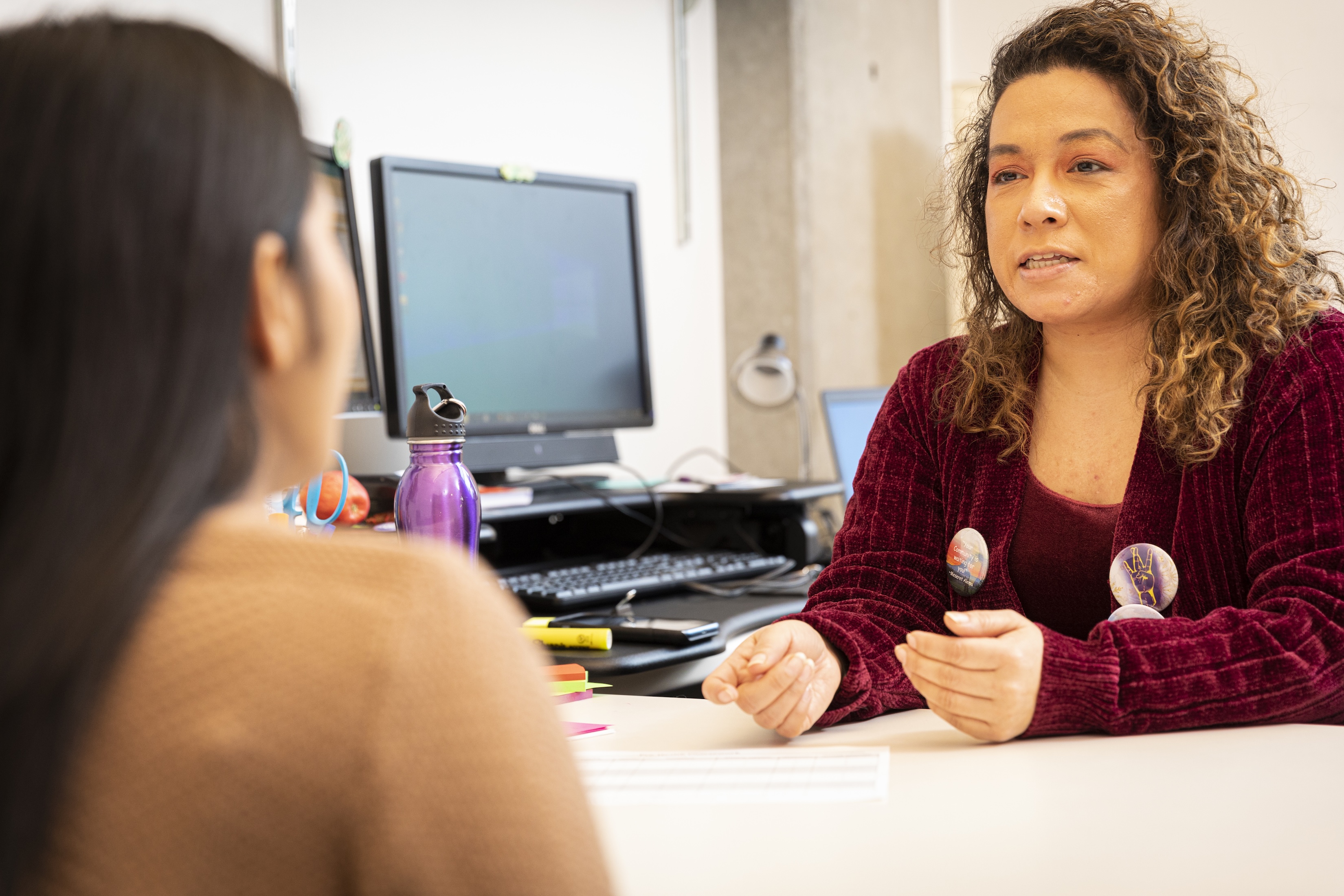Person with curly hair and a jacket seated speaking to another person with long hair.