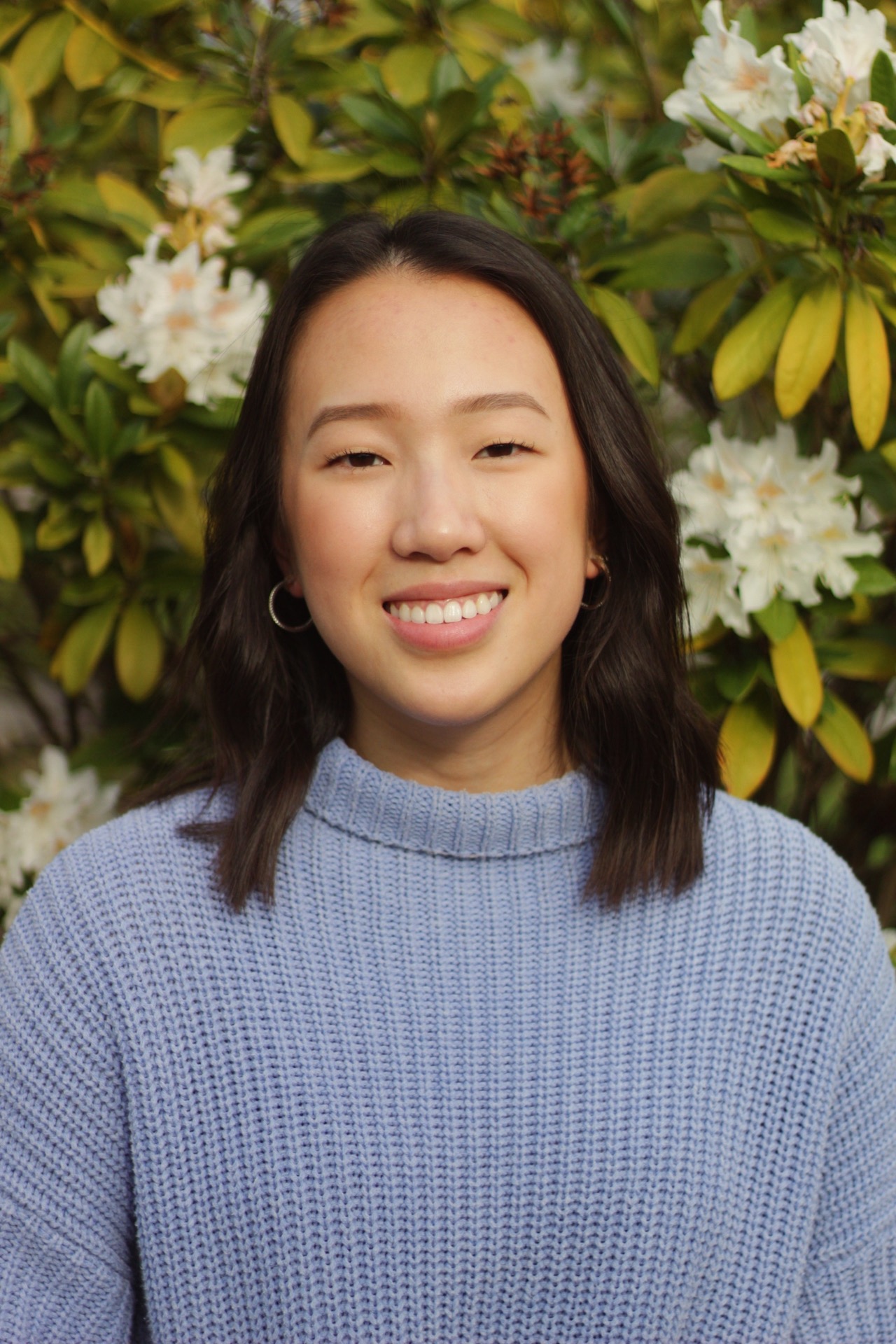 Patrice stands in front of a flowering bush and is wearing a light purple knitted sweater.