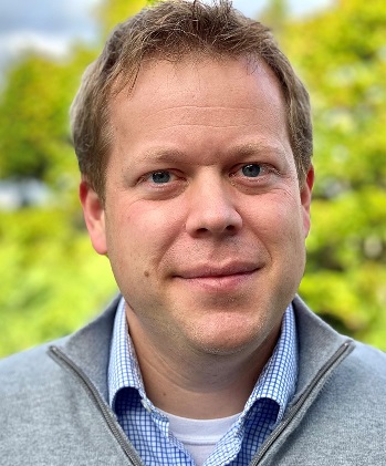 Danny looks at the camera wearing a grey sweater and blue collared shirt.